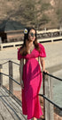 Woman wearing a bright pink Roma Dress with puffy sleeves and a fitted waist standing outdoors by a wooden railing in a sunny setting