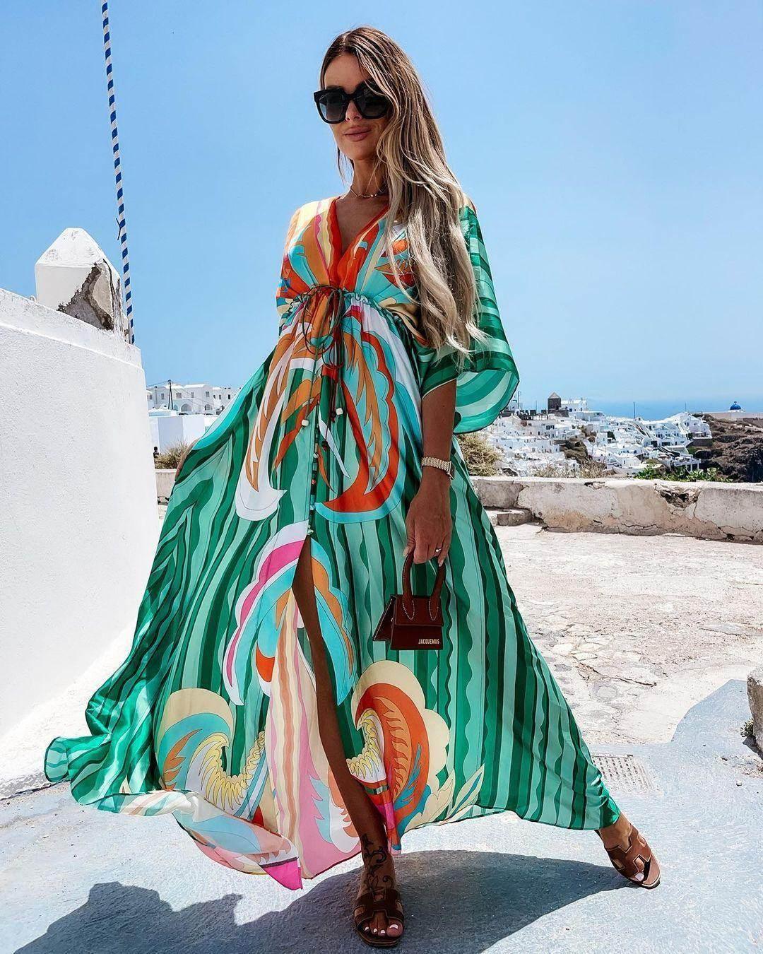 Woman wearing a flowing green Marcel Dress with colorful patterns and kaftan sleeves, standing outdoors with white buildings and a clear sky in the background