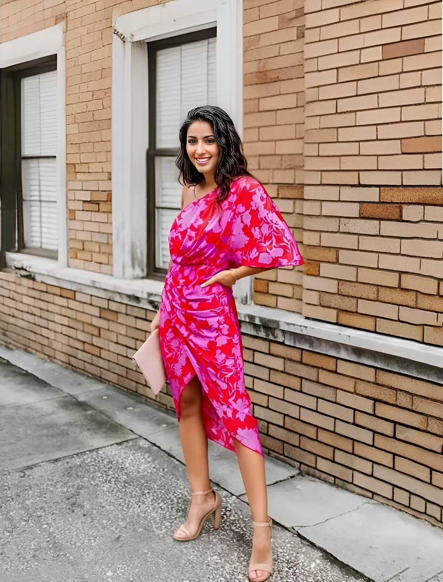 Woman wearing a stylish pink floral Razi Dress with one-shoulder design and slit, standing by a brick wall outdoors